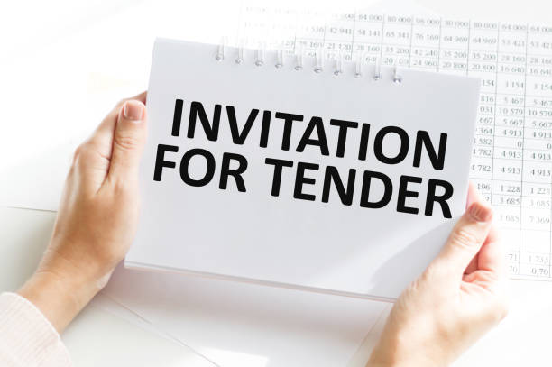 istock INVITATION FOR TENDER, text on white notepad in the hands of a businessman on a table next to financial reports 1403237181
