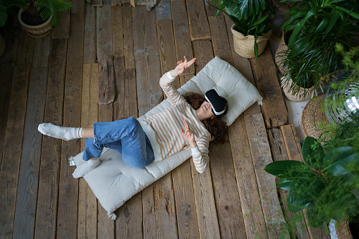 Young astonished woman in casual clothes lying on floor in greenhouse feeling excited while using 360 VR headset, exploring augmented reality at home, immersive technology virtual reality concept