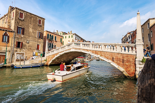 Venice, Italy - Sept 13, 2015: Ponte delle Guglie (bridge of the spires - 1580) over the Canale di Cannaregio (Cannaregio canal) of the Venetian lagoon. Venice, UNESCO world heritage site, Italy, Europe. A group of tourists cross the ancient bridge on a sunny summer day, while other people travel along the canal aboard small water taxi.