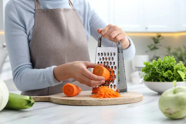 Woman grating fresh ripe carrot at kitchen table, closeup