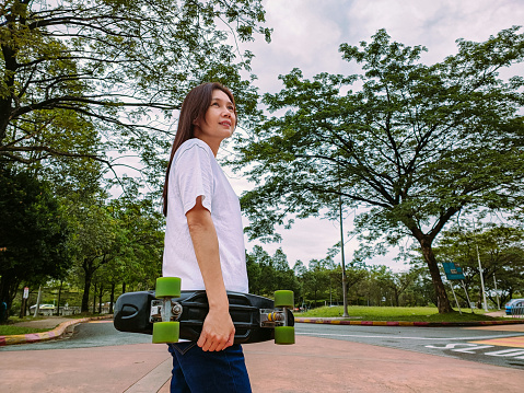 Portrait of female skateboarder in casual clothes on the empty road of the path near the parking area at twilight with her skateboard.