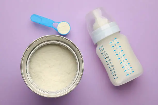 Photo of Feeding bottle with infant formula and powder on violet  background, flat lay