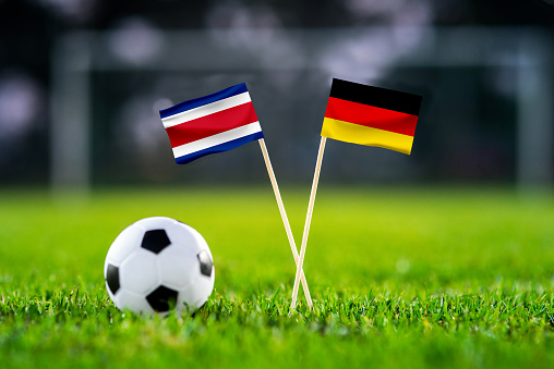 October 2022: Costa Rica vs Germany, Football match wallpaper, Handmade national flags and soccer ball on green grass. Football stadium in background. Black edit space.