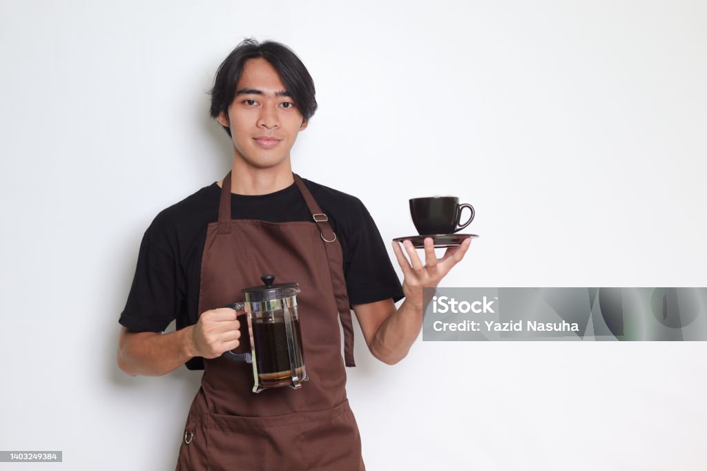 Asian barista man showing a cup while holding french press coffee maker Portrait of attractive Asian barista man in brown apron showing a cup and saucer while holding french press coffee maker. Isolated image on white background French Press Stock Photo