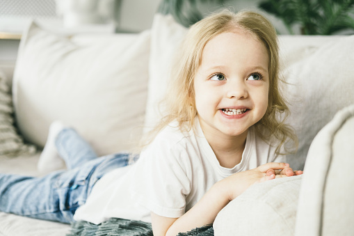 Little girl with blond hair having fun at home. Happy smile on the childs face. The child smiles mischievously. Close-up portrait of a 4 year old girl. Little girl lying on her stomach on the couch