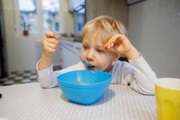 istock Blond toddler sitting at dining table and eating cereal from bowl, looking tired 1403252557