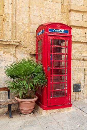 Mdina, Malta - Sep 26, 2021: Red telephone box to pay calls with a card next to a green plant in Mdina