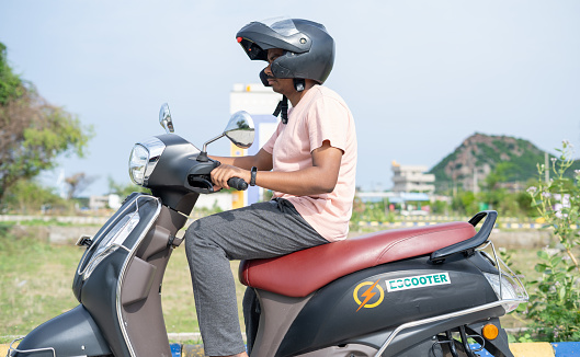 Man driving Ev or electric scooter by wearing helmet on road - concept of safety, technology and lifestyle