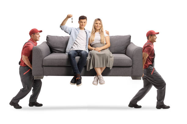 movers carrying a young couple sitting on a gray sofa and holding a house key - real estate imagens e fotografias de stock