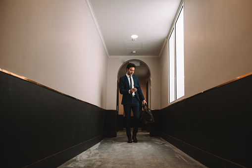 Businessman in a luxury hotel walking searching the room. Meanwhile is texting on mobile phone receiving a good news.