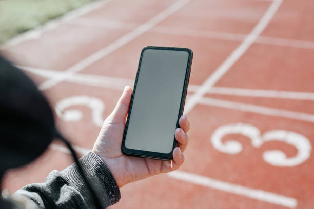 Sports and technology concept. Sportswoman using mobile phone with blank white screen while exercising in outdoor stadium. Close-up of woman's hand holding smartphone with an empty advertising space stock photo