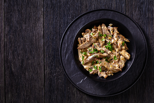 liver stroganoff with wholegrain mustard, onion and mushrooms in black bowl on dark wooden table, horizontal view from above, flat lay, free space