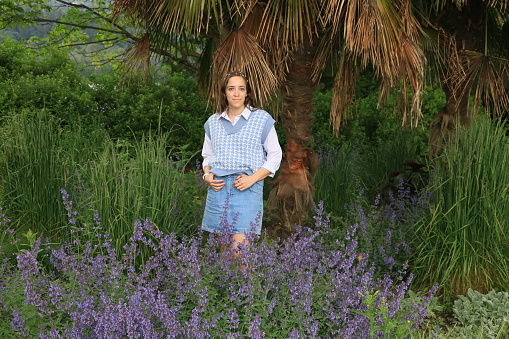 A Mexican woman standing in a garden in Spring. She is wearing A white shirt, blue pattern sleeveless sweater, denim skirt, and a large watch.