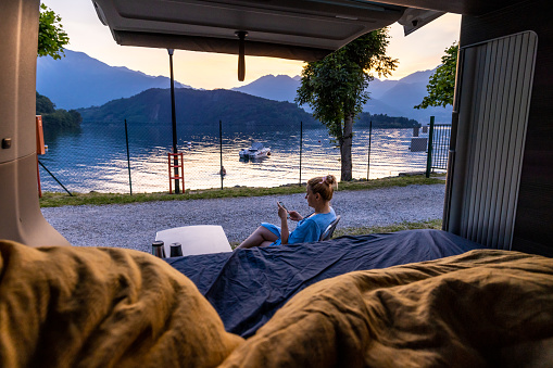 Woman sitting on chair text messaging on smart phone next to parked motor home while camping by Lake Como