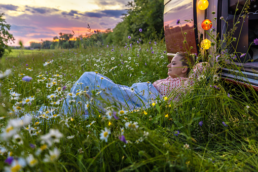 Carefree woman relaxing over grass amidst flowering plants outside camper trailer against sky during sunset
