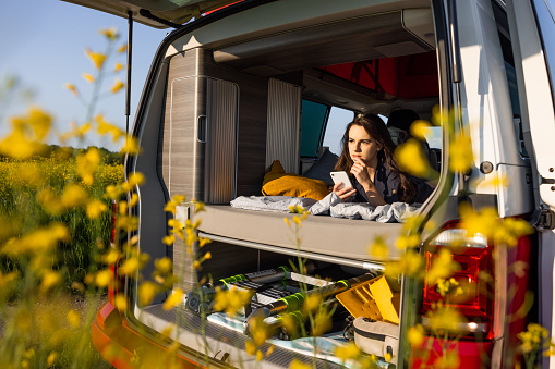 Young adult woman with long brown hair lying down in her van,back door of van is opened,concentrated facial expression,holding a smartphone,yellow flowers in front,van life
