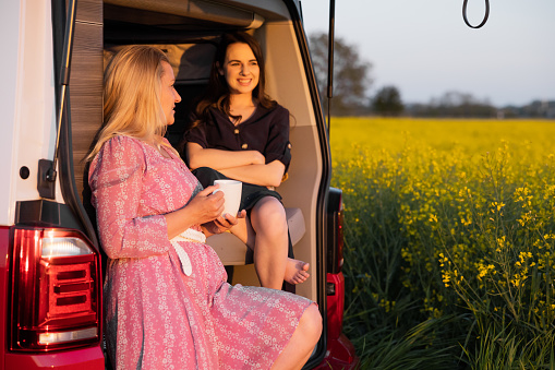 Mother with long blond hair and daughter with long brown hair relaxing,sitting at the trunk of their van,smiling and talking,mother is holding a cup,during sunset,van life