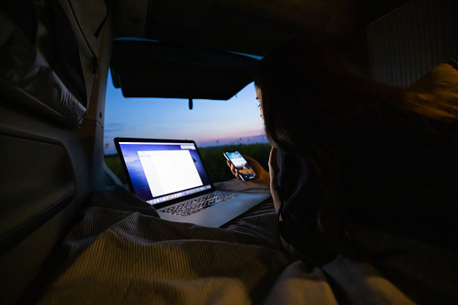 Young adult woman with long brown hair lying down in her van and working on her laptop,holding a smartphone,in the evening,doors of van are opened,van life