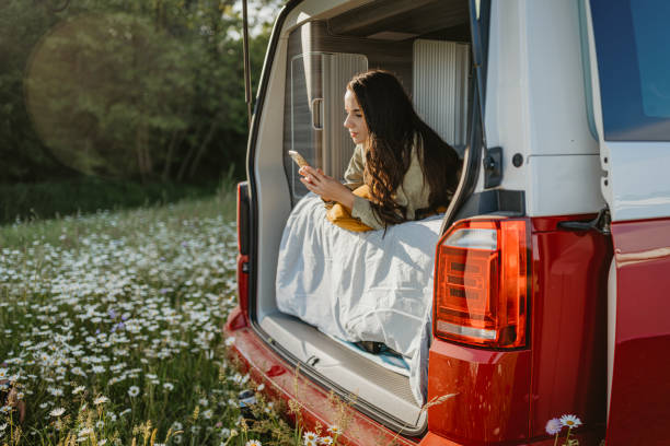 Woman with brown hair relaxing and lying down in her van with opened back door, looking down at her smartphone Woman with long brown hair relaxing and lying down in her van with opened back door, looking down at her smartphone, van life in Jeruzalem Iphone stock pictures, royalty-free photos & images