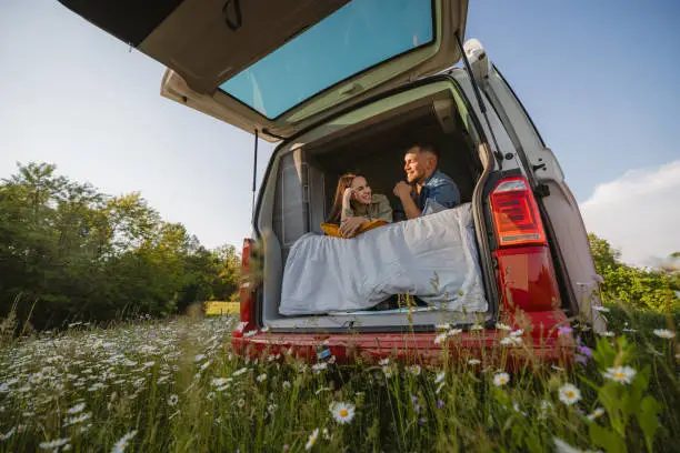 Photo of Couple smiling at each other, lying down on a mattress in their van, back door of van is opened, parked on a meadow in the nature