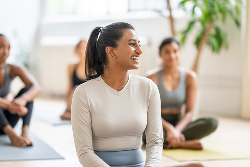 A small group of adults sit around on yoga mats as they take a break between fitness classes. They are each dressed comfortably in athletic wear and are talking amongst each other.