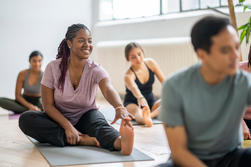 A group of adults sit individually on yoga mats spread around a studio as they warm up their muscles before class.  They are each dresed comfortably in athletic wear and are smiling as they stretch out their legs.