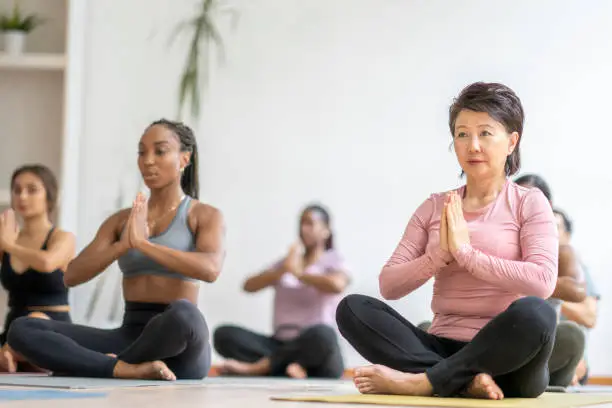 A large group of diverse adults participate together in a meditation class.  They are each seated on a yoga mat with their legs crossed and their hands in a prayer pose as they focus on their breathing.
