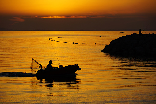 A fisherman with a fishing rod in his hand and a fish caught stands in the water against a beautiful sunset.