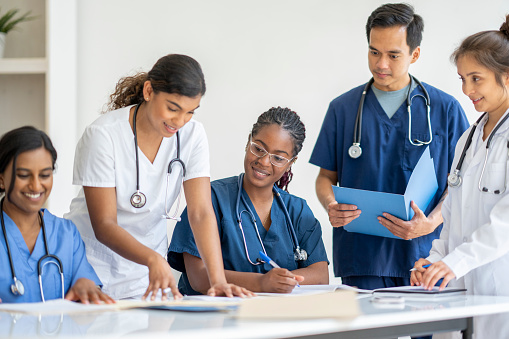 A small group of medical students sit around a table as they study together.  They are each dressed in medical scrubs and have various reference materials and file folders scattered on the table in front of them as they work together.