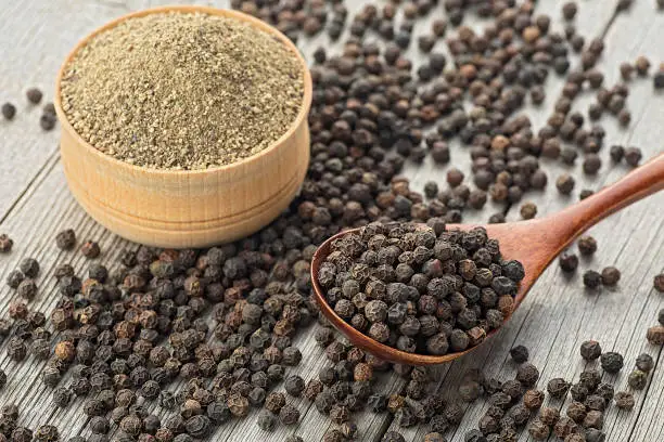 Photo of Black peppercorns and ground black peppercorns on wooden table. Milled black pepper in wooden bowl. Black peppercorns in wooden spoon.