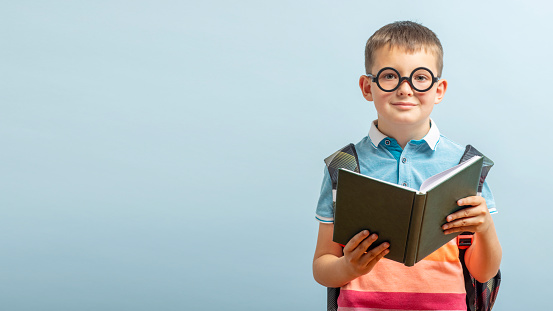 Little schoolboy in eyeglasses is reading a book on blue background. Education and school concept. Mockup, copy space