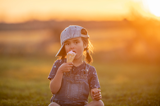 A sweet little brunette hired girl with pigtails sits in the grass on a hot summer evening as she enjoys an ice cream cone.  She is dressed in a denim jumper and looking at the camera with a neutral expression as she licks her cool summer treat.