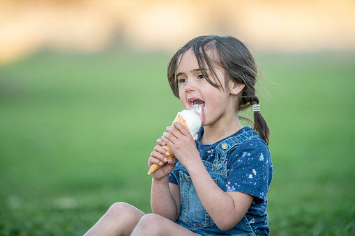 A sweet little brunette hired girl with pigtails sits in the grass on a hot summers day as she enjoys an ice cream cone.  She is dressed in a denim jumper and smiling as she licks her cool summer treat.