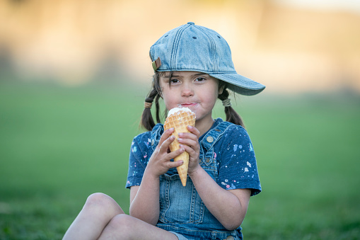 A sweet little brunette hired girl with pigtails sits in the grass on a hot summers day as she enjoys an ice cream cone.  She is dressed in a denim jumper and smiling as she licks her cool summer treat.