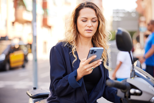 A woman sits on a scooter and looks at a phone screen.
