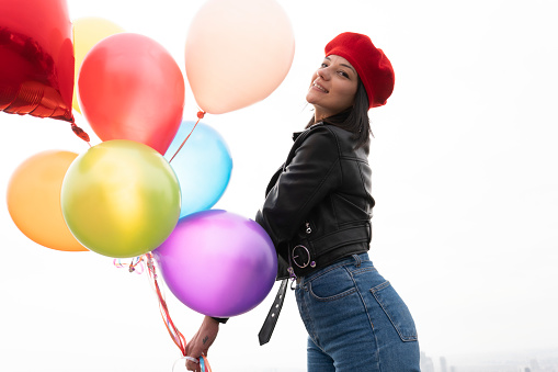 Happy woman holding multicolored balloons in the air, and looking at them joyfully. She is standing in front of a blurred background where a bit of city and a massive piece of sky are to see.