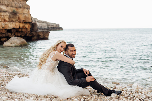 Married couple in wedding dress and official suit sitting on the coast of the sea or ocean near cliff rocks mountains. Honeymoon abroad in Europe, Italy. Embracing each other, love bounding. Event