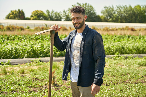 Three-quarter front view of Buenos Aires man in work clothes standing outdoors in herb garden, holding long handled tool, and smiling at camera.