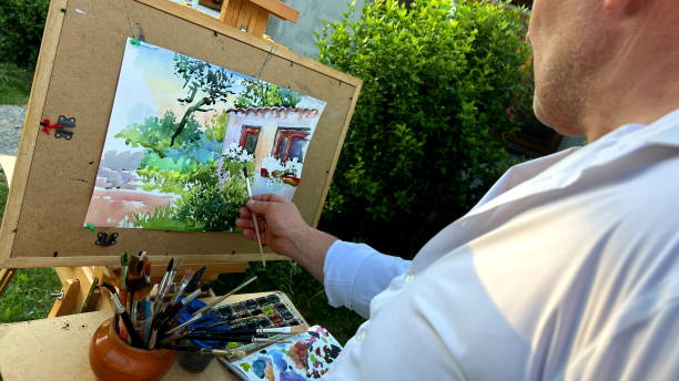 artista masculino adulto de 45 a 55 anos de idade pinta um quadro olhando para sua casa na frente dele uma folha de papel em que você já pode ver as janelas e madeira verde, bem como muitas tintas e pincéis ele está em uma camisa branca e na frente del - male 45 years old - fotografias e filmes do acervo