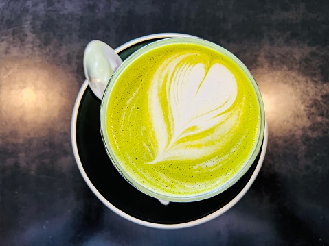 Horizontal high angle closeup photo of a cup of green matcha latte with a heart froth art pattern on a round black saucer with a teaspoon, on a shiny black table in a cafe.