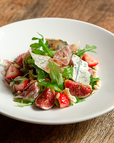 Salad with hamon ham, figs, blue cheese, arugula and strawberry on a plate. Dark Wooden background.