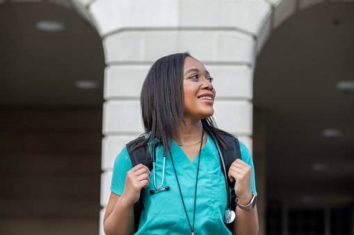 Young African American woman smiles while walking on campus of medical school or nursing college. Intern or student learning a healthcare occupation is wearing medical scrubs and a stethoscope, and is carrying a backpack. Woman is outdoors near educational buildings
