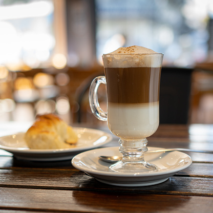 a cappuccino with a croissant on a wooden table in a bar. the cappuccino is in a glass cup, you can see the two colors and the foam.