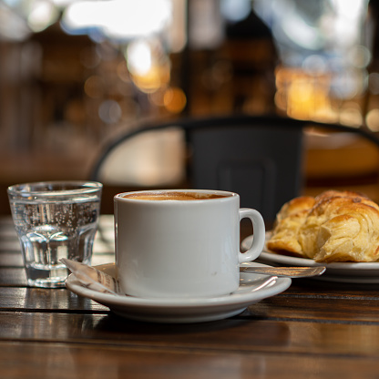 a cup with coffee with milk, a croissant and a glass with water on a wooden table in a bar.