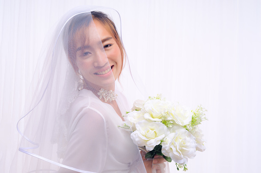 studio portrait of asian bride in white gown with veil holding flower bouquet on white background