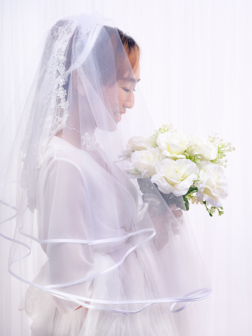 studio portrait of asian bride in white gown with veil holding flower bouquet on white background