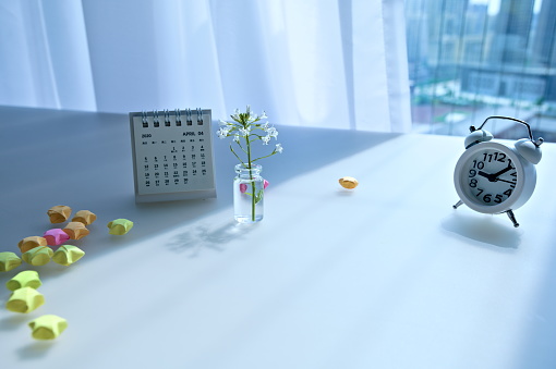 Modern home office desk with alarm clock, calendar, flowers in glass bottle with water, and colorful paper stars decor. Interior minimal table near window and curtain