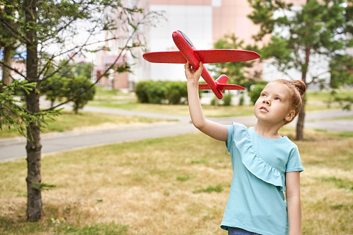 Little girl with airplane toy. Adventure fly concept. Stay home game. Family garden activity. Red hair female portrait. Styrofoam childhood pilot. Summer time. Park outdoor aviation
