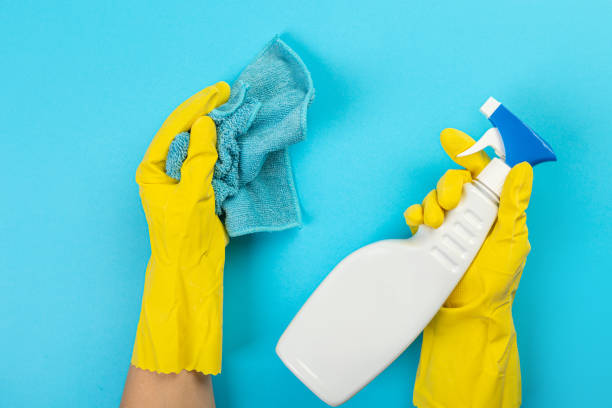 The hands of a housewife in yellow rubber protective gloves hold a bottle of household chemicals and a rag. Detergent for various surfaces in the kitchen, bathroom and other rooms. Light pastel blue background. las manos de un ama de casa con guantes protectores de goma amarilla sostienen una botella de productos químicos domésticos y un trapo. detergente para diversas superficies en la cocina, baño y otras habitaciones. fondo azul pastel claro.