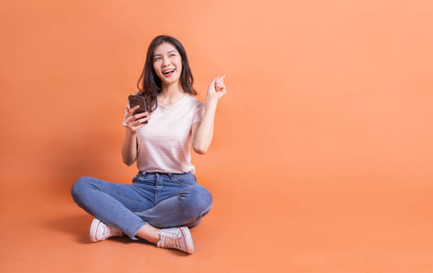 immagine a figura intera di una giovane ragazza asiatica che utilizza lo smartphone su sfondo arancione - filipino ethnicity immagine foto e immagini stock